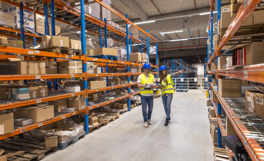 Two caucasian warehouse workers walking in distribution storage area discussing about logistics and organization.